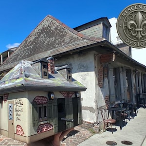 May include: A miniature replica of Lafitte's Blacksmith Shop Bar, featuring a gray roof, beige walls with red brick accents, green shutters, and a small entrance door. The text on the side reads Lafitte's Blacksmith Shop Bar in black script.