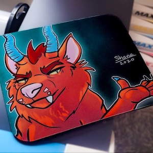 May include: A red and blue cartoon character with horns and a toothy grin. The character is giving a thumbs up. The background is dark blue. The image is signed "Shane 2020".