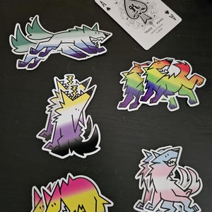 Peut inclure: Plusieurs autocollants de loup colorés avec des drapeaux de la fierté différents. Les autocollants présentent un loup courant, un loup hurlant et deux loups enlacés. Une carte à jouer avec un motif de pique est également présente.