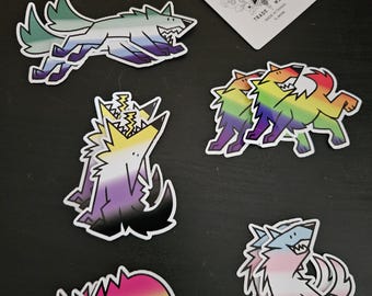 May include: A collection of colorful wolf stickers with various pride flag designs. The stickers feature a running wolf, a howling wolf, and two wolves embracing. A playing card with a spade design is also present.