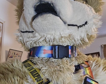 Könnte beinhalten: Ein pelziges braunes und weißes Hundekostüm mit einem schwarzen Halsband und einem blauen und orangefarbenen Knochenmuster. Das Kostüm hat auch ein gelbes und schwarzes Geschirr mit dem Text "Keep an eye on me" und ein regenbogenfarbenes Geschirr.
