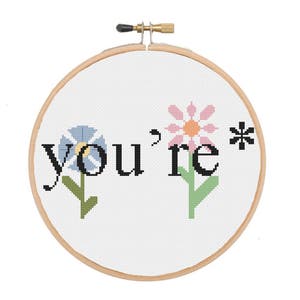 May include: A white embroidery hoop with the phrase "you're*" stitched in black thread. Two flowers, one blue and one pink, are stitched on either side of the phrase.