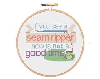 May include: A white embroidery hoop with a cross-stitch design that reads "If you see a seam ripper now is not a good time". A small green frog with a red tongue is sitting on a green leaf.
