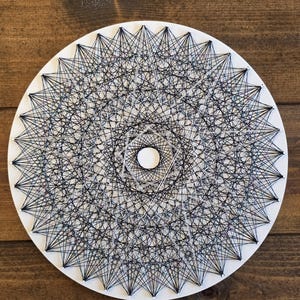 May include: A white, circular string art piece with a complex geometric design. Black and white thread is intricately woven to create a starburst pattern. The artwork is mounted on a wooden surface.