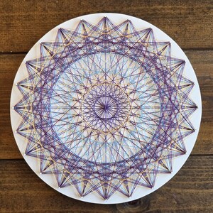 May include: A round, white string art piece with a geometric design. The artwork features a complex pattern of purple, gold, and light blue threads stretched across nails, creating a starburst effect. The piece is displayed on a wooden surface.