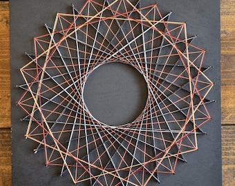 May include: Geometric string art on a black square. The design features a circular pattern with red, orange, white, and black threads. The artwork is mounted on a black surface, creating a striking contrast.