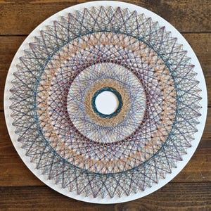 Könnte beinhalten: Runde String-Art-Arbeit mit weißem Hintergrund und komplexem geometrischem Design. Das Kunstwerk verwendet Fäden in Blau, Lila, Beige und Grün, um konzentrische Kreise zu bilden, ein dekoratives handgefertigtes Wandobjekt.