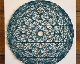 May include: A teal string art piece with a geometric design. The artwork is a circular shape with a complex web of teal thread stretched between small white pins. The piece is mounted on a white square, set against a wooden background.