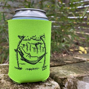 May include: A green can cooler featuring a black and white illustration of an alligator relaxing in a hammock. The alligator is in a hammock suspended between two trees. The text "Hangin' with the Gators" is printed on the cooler.