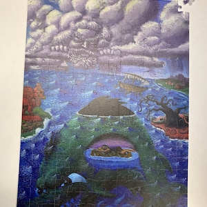 May include: A completed jigsaw puzzle showing a fantastical scene with a large green sea creature, a small island with a tree, and a stormy sky with a flock of birds. The puzzle is set against a blue background.