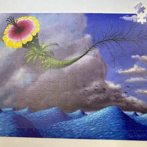 May include: A surrealist jigsaw puzzle featuring a green, leafy creature with a large pink and yellow flower. The creature is flying through a cloudy sky with a bare tree branch extending from its body. The sky is divided into two sections, one blue and one grey. The puzzle is missing one piece. The bottom of the image shows a blue ocean with waves.
