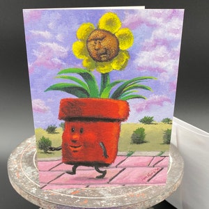 May include: Greeting card featuring an original painting of a walking flower pot with a sunflower head. The flower pot is red-orange with a smiling face and black feet. The sunflower has a face and is set against a sky with clouds.