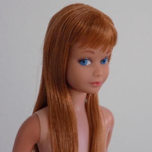 May include: A vintage doll with long, straight auburn hair and bangs. The doll has blue eyes, a fair complexion, and a slight smile. The doll is wearing no clothing and is set against a plain white background.