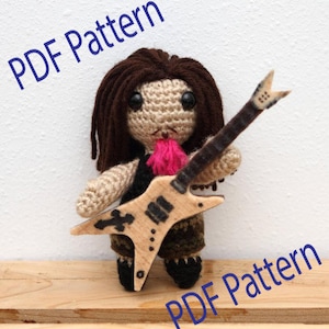 May include: A crocheted doll with brown hair and a black vest, holding a wooden guitar. The doll has a pink tongue and is wearing dark green pants and black shoes. The text "PDF Pattern" is written in blue.