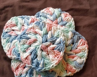 May include: Three crocheted coasters in a light blue, pink, and green color scheme. The coasters are made with a textured, circular design.