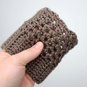 May include: A brown crocheted cup cozy with gold glitter yarn. The cozy is made with a simple stitch pattern and has a slightly textured look.