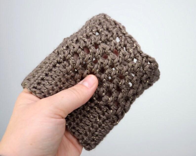 May include: A brown crocheted cup cozy with gold glitter yarn. The cozy is made with a simple stitch pattern and has a slightly textured look.