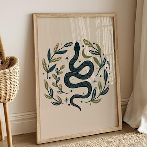 Op de afbeelding: Een ingelijste kunstprint met een donkergroene slang in een S-vorm, omringd door een cirkelvormige compositie van groene bladeren, kleine sterren en een maansikkel op een lichtbeige achtergrond, geplaatst in een lichthouten lijst.