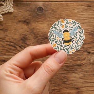 May include: A circular sticker featuring a central yellow and black striped bee with light blue patterned wings, surrounded by a decorative wreath of green leaves, small orange flowers, and thin stems on a cream-colored background.