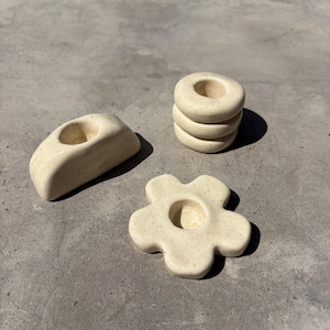 May include: Three off-white ceramic candle holders. One is a curved shape, another is a stack of three rings, and the third is a flower shape. All have speckled surfaces and a matte finish. The candle holders are on a grey surface.