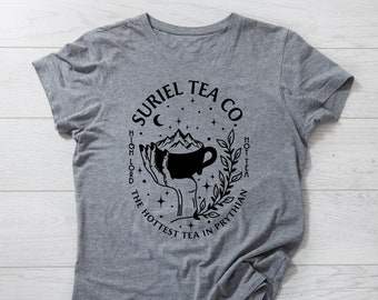 以下が含まれることがあります： 黒いグラフィックデザインが施されたグレーのTシャツ。手は、中に山が入ったティーカップを持っています。グラフィックの周りのテキストは、「Suriel Tea Co. High Lord The Hottest Tea In Prythian Hot Tea」です。
