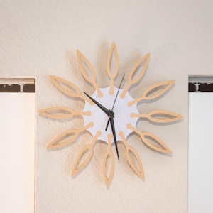 May include: A decorative wall clock with a sunburst design. The clock features a white face with black hands and a light brown wooden frame. The clock is mounted on a beige wall.