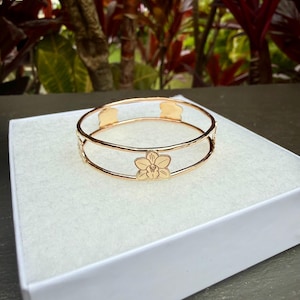 May include: Gold-colored bracelet with a floral design. The bracelet features a double-band design with a flower in the center and decorative cutouts. The bracelet is displayed on a white surface, with a blurred background of green and red foliage.