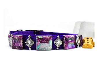 May include: A purple dog collar with square, abstract-patterned accents in shades of purple, pink, and teal. The collar features silver-toned diamond-shaped embellishments with clear rhinestones and silver studs. The collar is made of a purple fabric.