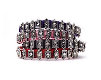 Könnte beinhalten: Gestapelte Armbänder mit silberfarbenen Metall- und Schmuckakzenten. Die Armbänder haben ein quadratisches Design mit einem zentralen Juwel und sind auf Samtbändern in Rot, Rosa und Lila angebracht.