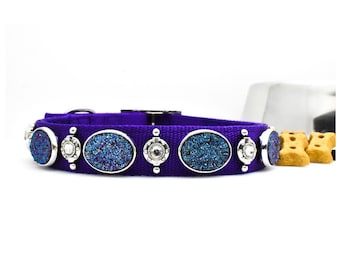 May include: A purple dog collar with silver-colored embellishments. The collar features oval-shaped, blue-glittered stones set in silver frames, alternating with smaller, round silver accents. The collar is designed for pets.