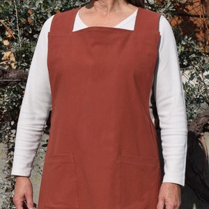 Puede incluir: Un delantal cruzado de color óxido con dos bolsillos grandes. El delantal está hecho de una tela resistente y tiene un diseño sencillo. El texto "Crossback Apron 1 5 Sizes" está impreso en la parte inferior del delantal. El texto "PDF SEWING PATTERN & TUTORIAL I BEGINNER +" está impreso debajo del nombre del delantal. El texto "DESIGN BY YARROW CREEK TM" está impreso en la parte inferior de la imagen.