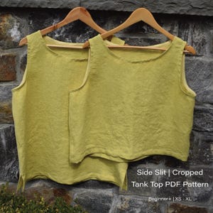 Pode incluir: Dois tops de tanque de linho amarelo com fendas laterais pendurados em cabides de madeira. Os tops são curtos e têm um caimento simples e relaxado. O texto "Side Slit | Cropped Tank Top PDF Pattern" está impresso abaixo dos tops. O texto "Beginner+ | XS - XL" está impresso abaixo do nome do padrão.
