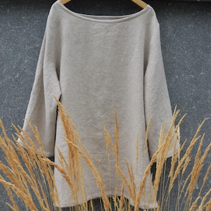 Peut inclure: Une tunique beige à manches longues avec une large encolure bateau, suspendue à un cintre en bois. Le vêtement présente une silhouette simple et ample avec des manches droites et un tissu texturé uni, présenté contre un mur gris foncé.