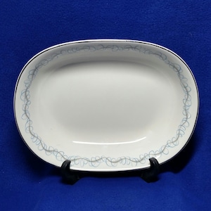 May include: White oval serving platter with a black rim and a light blue swirl design around the edge. The platter is displayed on a black stand against a blue background.