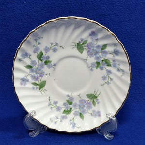 May include: White saucer with a gold rim and a floral pattern of blue and purple flowers with green leaves. The saucer has a ridged design and is displayed on a clear stand against a blue background.