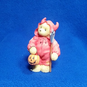 May include: A small, cream-colored bear figurine dressed in a red devil costume with a hood and horns. The bear holds a silver pitchfork and an orange jack-o'-lantern. The figurine is a Halloween-themed collectible.