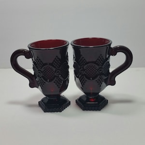 May include: Two ruby red vintage glass mugs with ornate detailing. Each mug features a pedestal base, a patterned body, and a curved handle. The interior of the mugs is also red. The mugs are likely for serving beverages.
