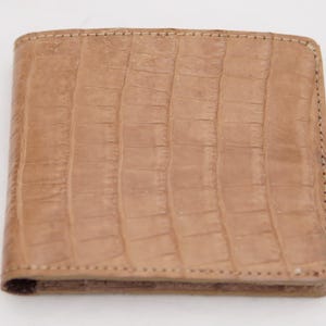 May include: A tan bi-fold wallet with a textured, crocodile-like pattern. The wallet is rectangular with rounded corners and a stitched border. The leather appears to be a light brown color.