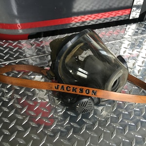 May include: A black firefighter's mask with a clear visor and a brown leather strap. The strap is personalized with the name "JACKSON".