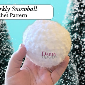 May include: A white crocheted snowball with a sparkly texture. The snowball is being held in a hand against a blue background with two small, snowy evergreen trees.