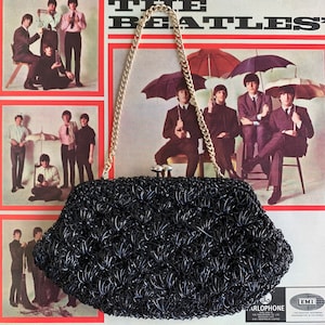 May include: A black crocheted evening bag with a gold chain strap. The bag has a clasp closure and is decorated with a small silver charm. The bag is photographed in front of a vintage Beatles record album.