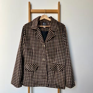 May include: A black and tan houndstooth blazer hangs on a wooden hanger. The jacket features a notched collar, two front pockets, and gold-coloured buttons. The blazer is displayed on a wooden ladder against a white wall.