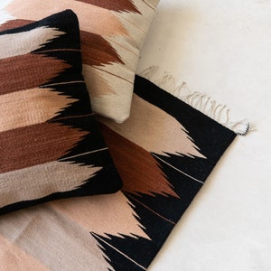 May include: A set of three woven pillows and a rug with a geometric pattern in shades of brown, black, and beige. The pillows and rug have a fringe detail.