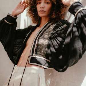 May include: A woman with curly brown hair wears a black cropped jacket with a white and black trim. The jacket has a V-neck and long sleeves. She is wearing a pair of light beige shorts.