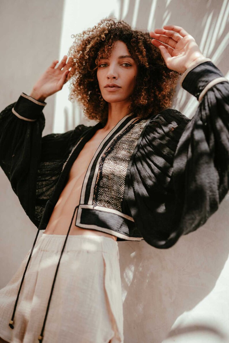 May include: A woman with curly brown hair wears a black cropped jacket with a white and black trim. The jacket has a V-neck and long sleeves. She is wearing a pair of light beige shorts.