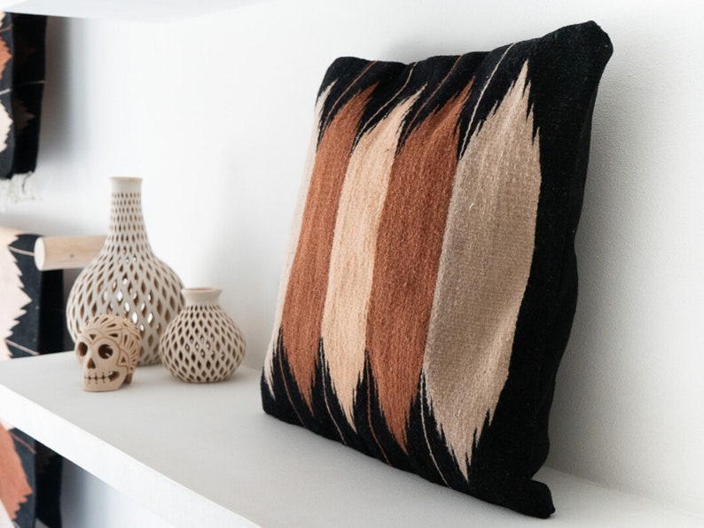 May include: A black, brown, and beige patterned throw pillow with a geometric design. The pillow is sitting on a white shelf with two decorative ceramic vases and a skull.