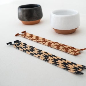 May include: Two woven bracelets with a houndstooth pattern. One bracelet is black and beige, and the other is brown and beige.