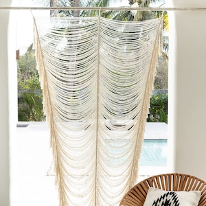 May include: A white macrame wall hanging with long fringe, hanging in front of a window with a view of palm trees and a swimming pool. A woven chair with a black and white patterned cushion sits in front of the wall hanging.