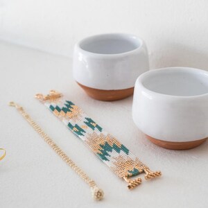 May include: Two white ceramic bowls with terracotta bases, a gold chain bracelet, and a beaded bracelet with a green, white, and gold geometric pattern.