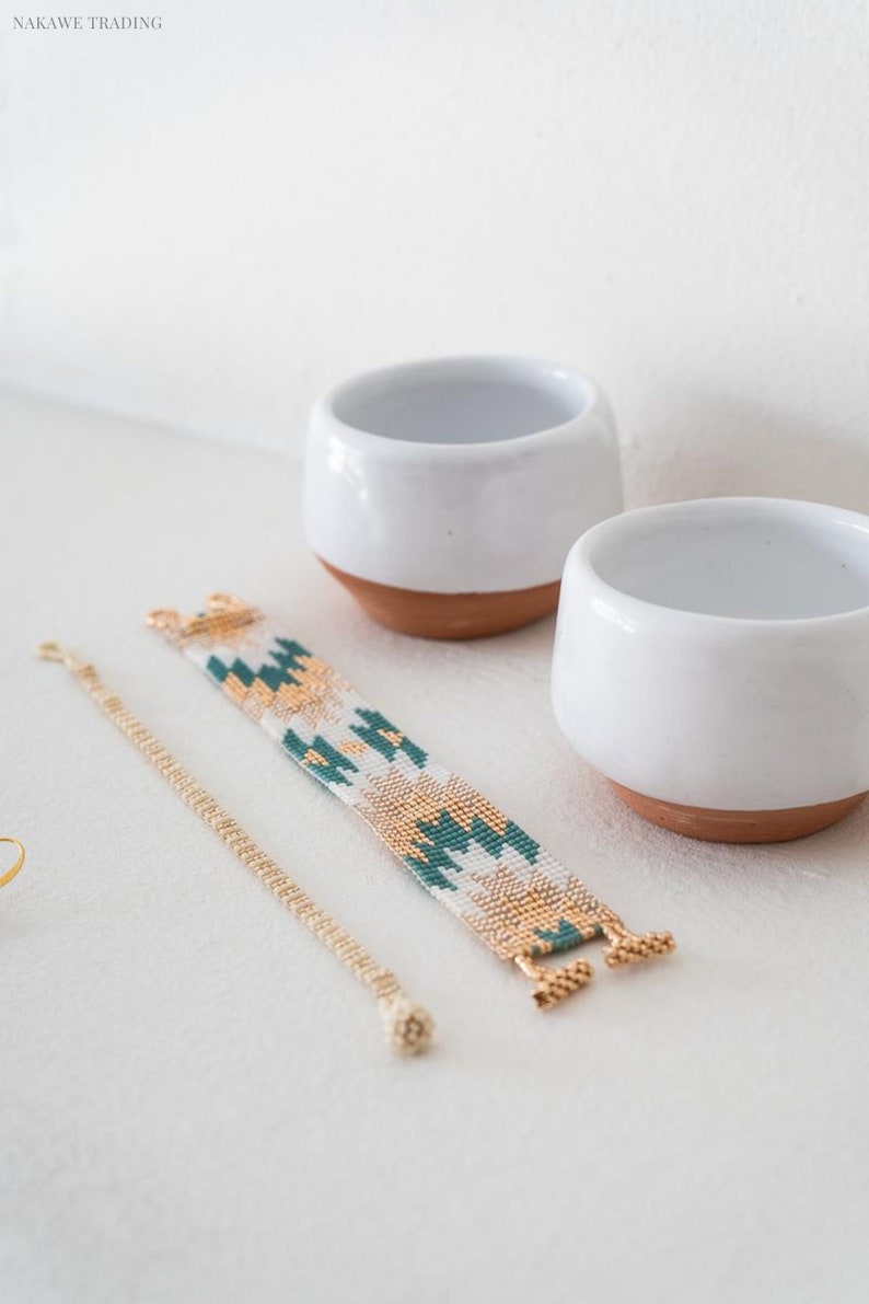 May include: Two white ceramic bowls with terracotta bases, a gold chain bracelet, and a beaded bracelet with a green, white, and gold geometric pattern.